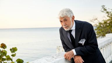Der jamaikanische Jazzpianist Monty Alexander in mediterranem Flair, auf eine Balustrade gestützt, im Anzug. Dahinter das Meer, er schaut etwas verschmitzt-fragend in die Kamera.