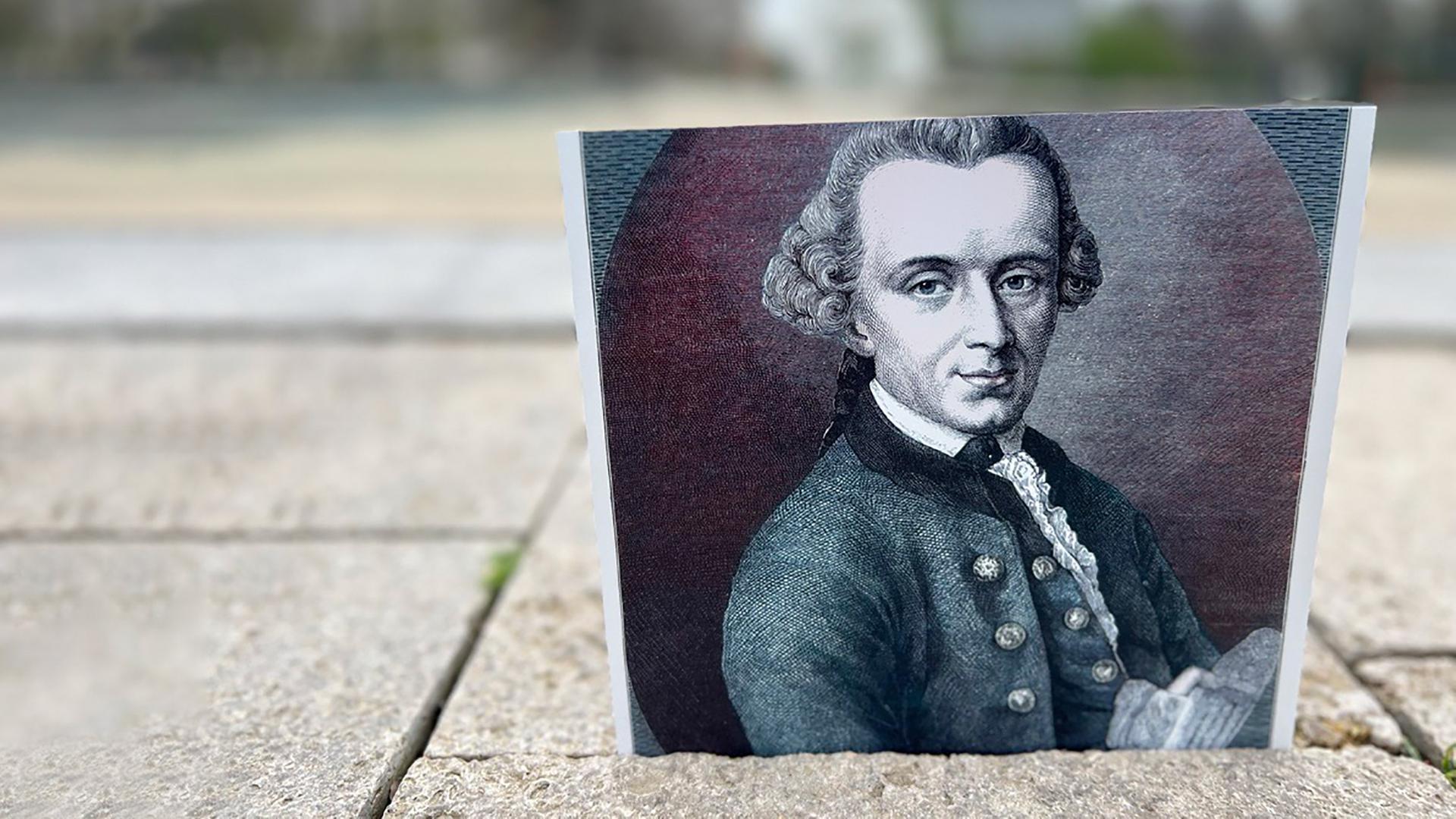 Auf einer gepflasterten Straße steht das schwarz-weiß-Foto von einem historischen Kupferstich, der einen Mann mit hoher Stirn und in Locken gelegten Haaren zeigt.