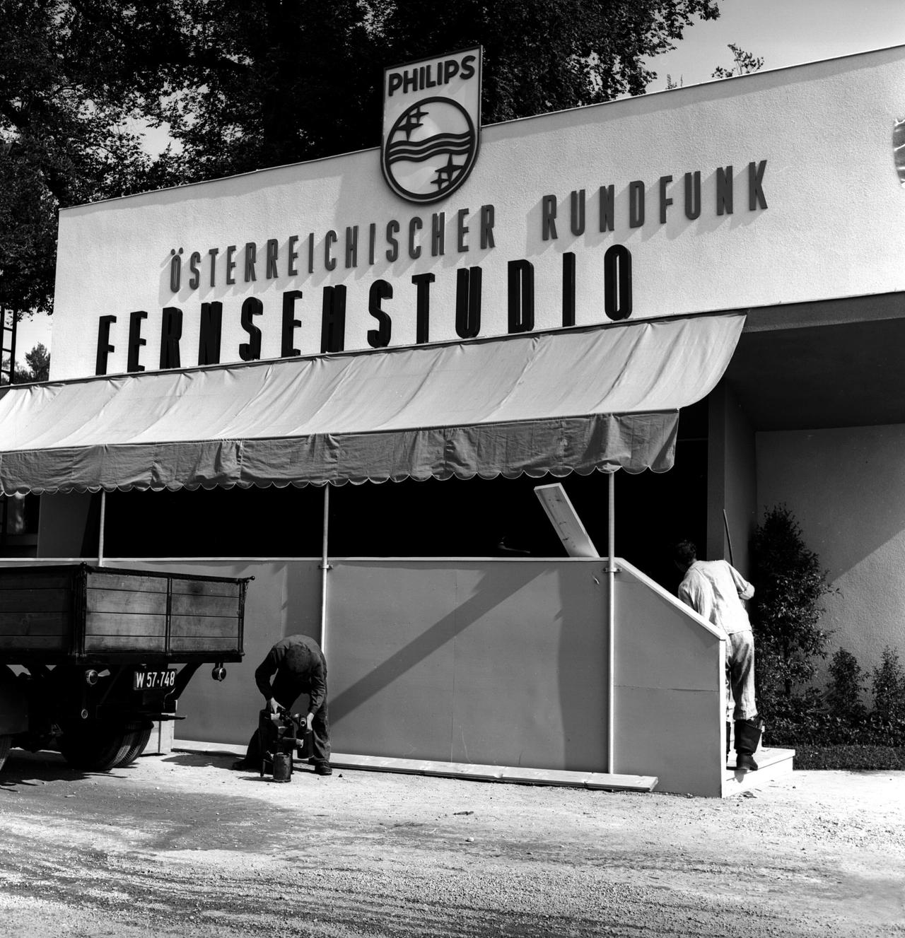Außenansicht eines Fernsehstudios des Österreichischen Rundfunks mit Philips-Logo im Jahr 1955, davor arbeiten zwei Männer.