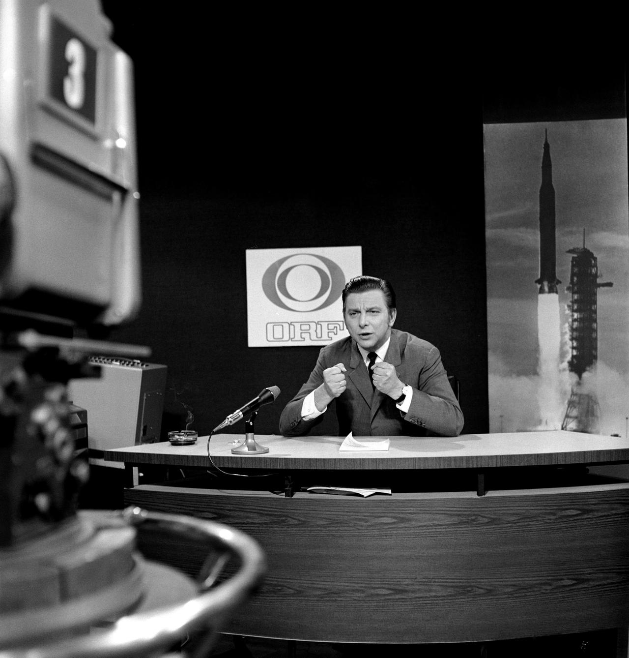 Dr. Hugo Portisch berichtet im ORF-Sonderstudio 1969 live über die Mondlandung von Apollo 11, im Hintergrund ein Bild einer startenden Rakete.