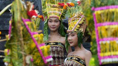 Zwei junge asiatisch aussehende Frauen tragen traditionellen balinesischen Kopfschmuck aus Blumen und Blättern und bestickte schulterfreie Wickelkleider.