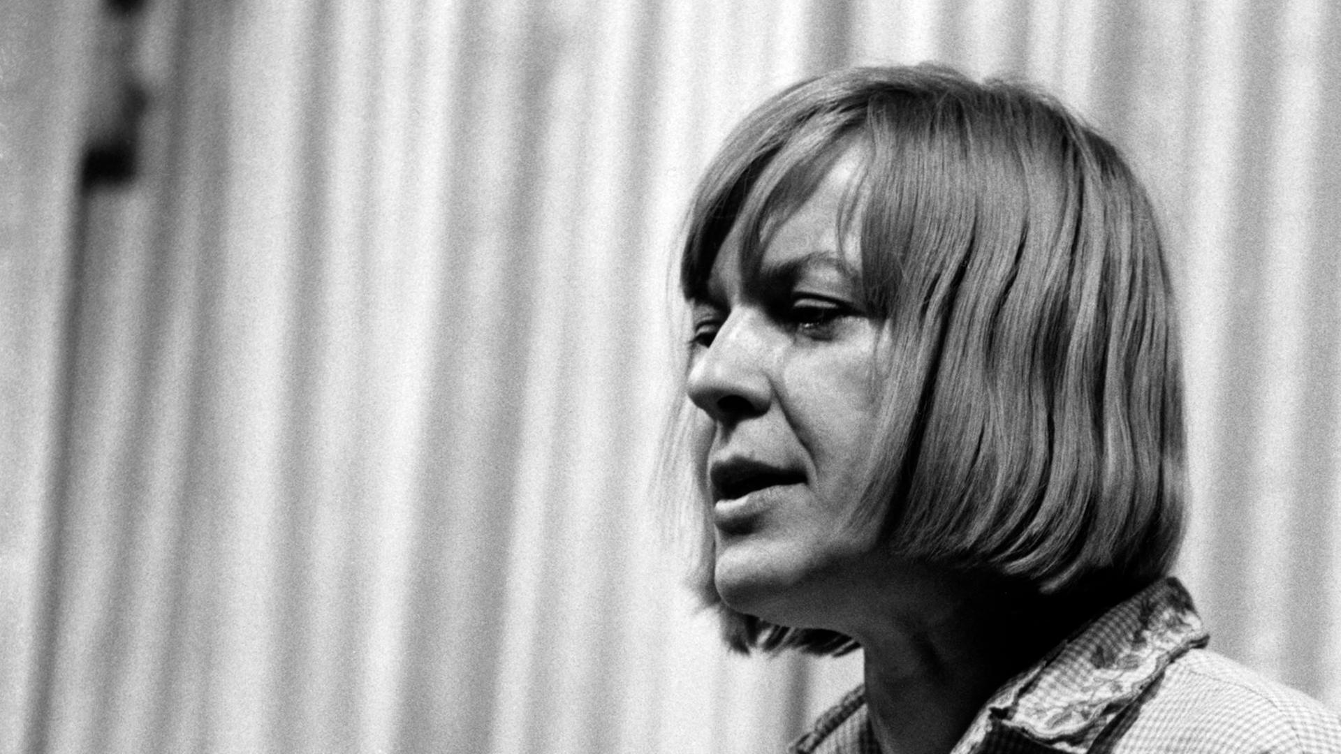 Altes Schwarz-Weiß-Foto einer Frau mit kinnlangen mittelblonden Haaren, die an einem Pult steht und etwas vorliest, oder redet.