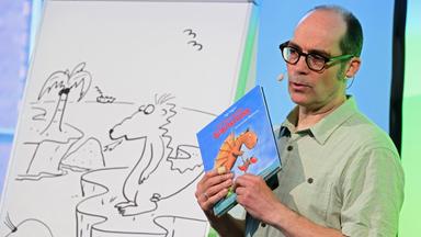 Links steht ein Whiteboard, auf das ein Drache, eine Palme und Vögel gemalt wurden. Daneben steht ein Mann, der ein Buch in der Hand hält, auf dem auch ein Drache zu sehen ist. 