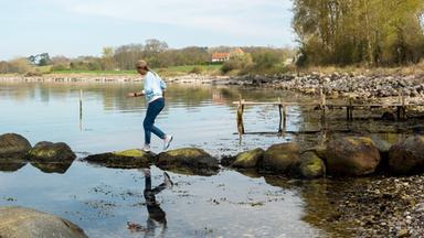 Eine blonde Frau mit Haarzopf läuft über große Steine in Richtung Wasser. Im Hintergrund ist das Ufer mit Wiesen, Bäumen und Häusern zu erkennen. Im Wasser schwimmt eine rote Boje.