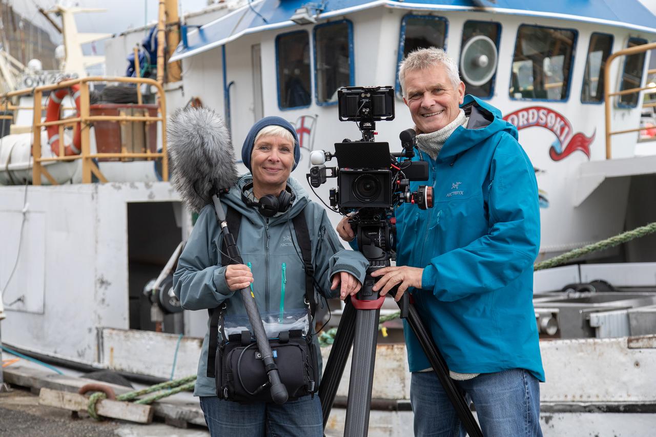 Dörte Hansen und Sven Jaax stehen vor einem Fischerboot. Sie hält ein Mikrofon mit Windschutz in der Hand. Er steht an der auf einem Stativ befindlichen Kamera.