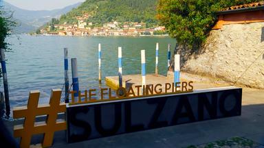 Ein Anlegeplatzan einem See, davor ein Schild mit „#The Floating Piers SULZANO“.