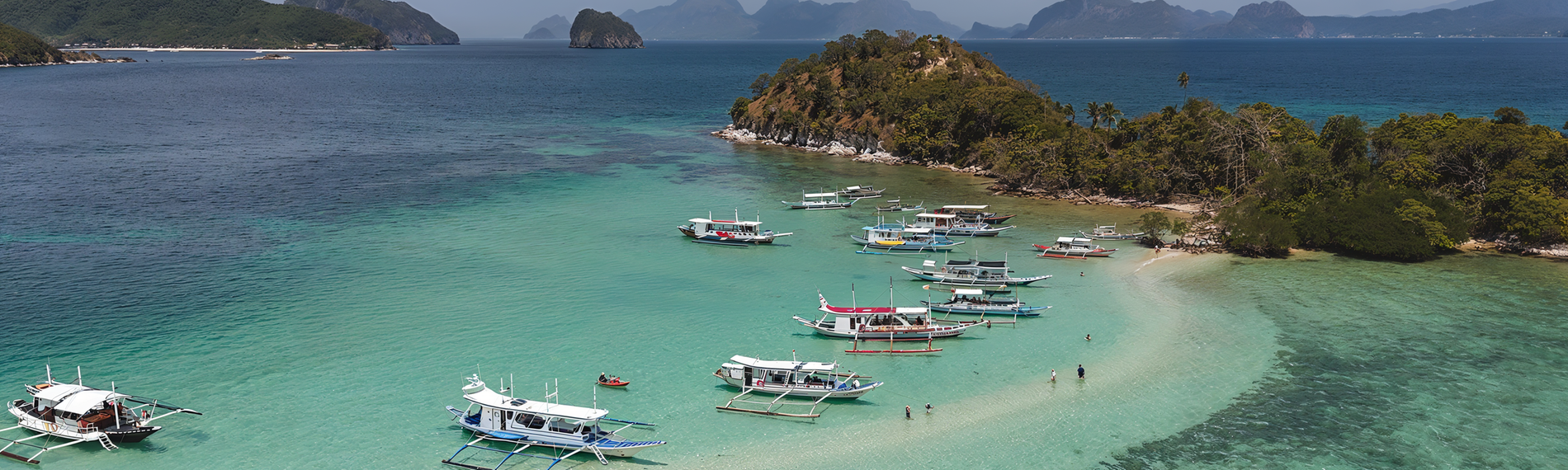 Schlangeninsel mit sandiger Landzunge vor der Küste von El Nido auf Palawan, umgeben von türkisfarbenem Wasser und traditionellen Auslegerbooten, mit bewaldeter Hügelinsel im Hintergrund.