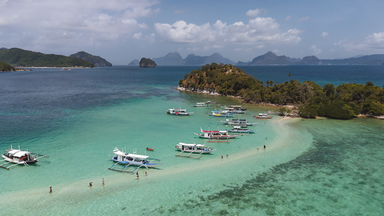 Schlangeninsel mit sandiger Landzunge vor der Küste von El Nido auf Palawan, umgeben von türkisfarbenem Wasser und traditionellen Auslegerbooten, mit bewaldeter Hügelinsel im Hintergrund.