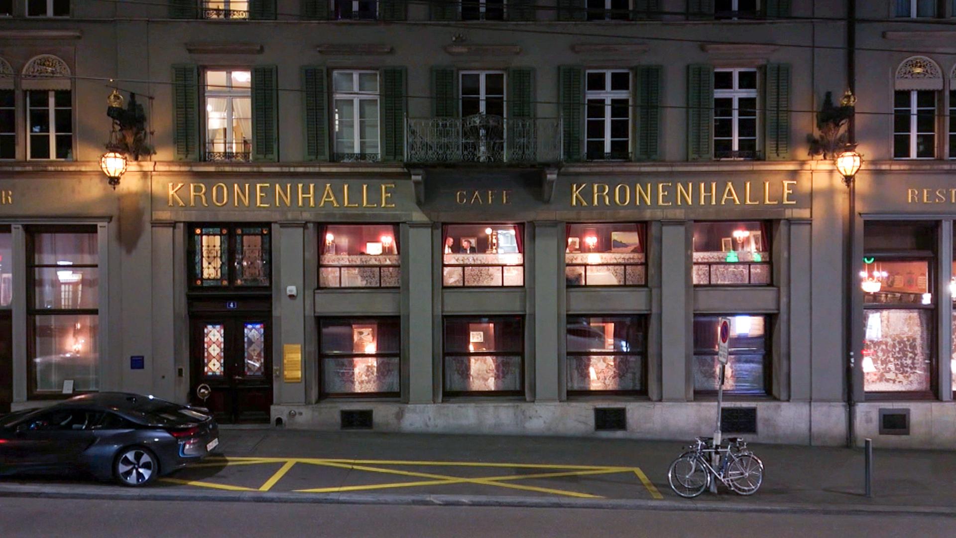 Außenansicht des traditionsreichen Restaurants Kronenhalle in Zürich bei Nacht, beleuchtete Fenster und Schriftzug gut sichtbar, davor ein geparktes Auto und zwei Fahrräder.