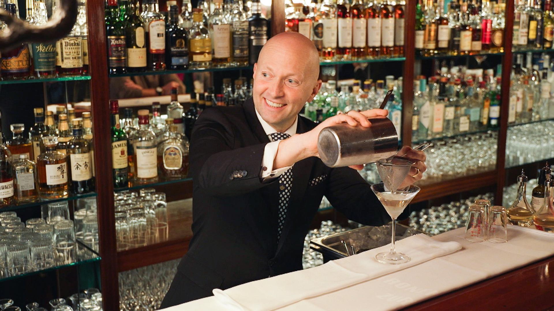 Christian Heiss, Chef de Bar in der Kronenhalle-Bar, mixt lächelnd einen Drink vor einer umfangreichen Sammlung edler Spirituosenflaschen.