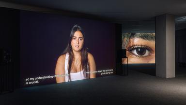 In einem dunklen Raum wird das Bild von einer jungen Frau an die Wand projiziert, in einem Nebenraum sieht man ein Großbild von ihrem Auge.