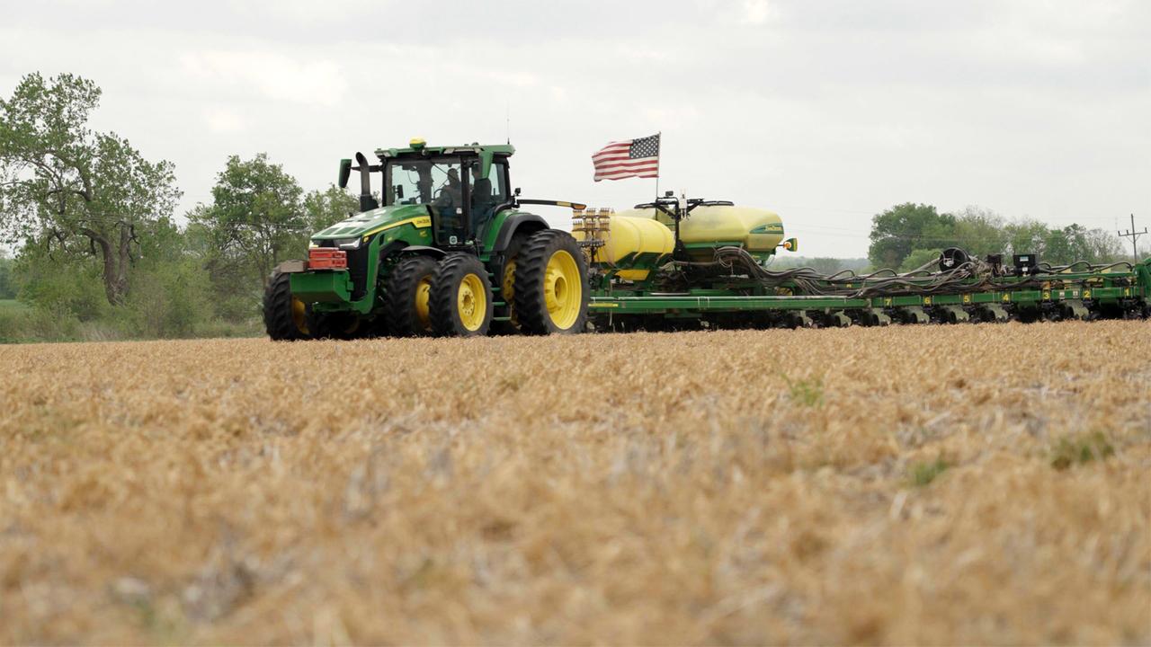 Das Bild zeigt einen landwirtschaftlichen Traktor, der auf einem trockenen Feld fährt. Der Traktor ist grün und hat gelbe Räder, und auf seinem Anhänger befindet sich eine gelbe Saatmaschine. Es weht eine US-amerikanische Flagge, die an dem Traktor befestigt ist. Im Hintergrund sind einige Bäume und eine lineare Struktur, möglicherweise Stromleitungen, sichtbar. Der Himmel ist bewölkt, was auf eine mögliche Wetterveränderung hinweist. Das Feld ist größtenteils braun und trocken, was auf eine Erntezeit oder Trockenheit hindeuten könnte.