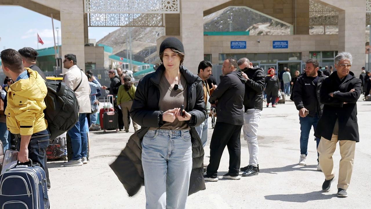 Das Bild zeigt eine belebte Szene an einem Grenzübergang zwischen Iran und der Türkei. Im Vordergrund steht eine Frau mit kurzer, dunkelblonder Haarfarbe, die mit einem dunklen Mantel und einer schwarzen Mütze bekleidet ist. Sie hält ein Mobiltelefon in ihren Händen und blickt in die Kamera.  Um sie herum sind viele Menschen zu sehen, die mit Gepäck, darunter Koffer und Taschen, unterwegs sind. Die Mehrheit der Personen trägt dunkle Kleidung. Einige Menschen stehen in Gruppen zusammen und scheinen zu unterhalten. Hinter ihnen befinden sich Gebäude mit Schildern, die auf den Grenzübergang hinweisen.  Im Hintergrund sind Berge sichtbar, die die Struktur der Landschaft betonen. Es herrscht viel Bewegung und reges Treiben, was auf eine geschäftige Grenzstation hindeutet.  Die gesamte Szene vermittelt ein Gefühl von Unsicherheit und hektischem Treiben, was durch die aktuellen politischen Ereignisse verstärkt wird.