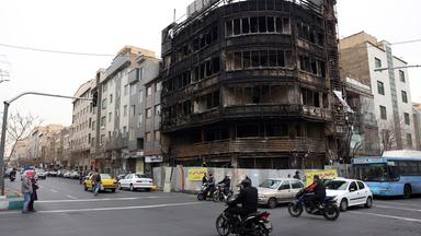 Über eine Straßenkreuzung an der ein ausgebranntes Geschäftshaus steht, fahren Motorradfahrer und Autos.