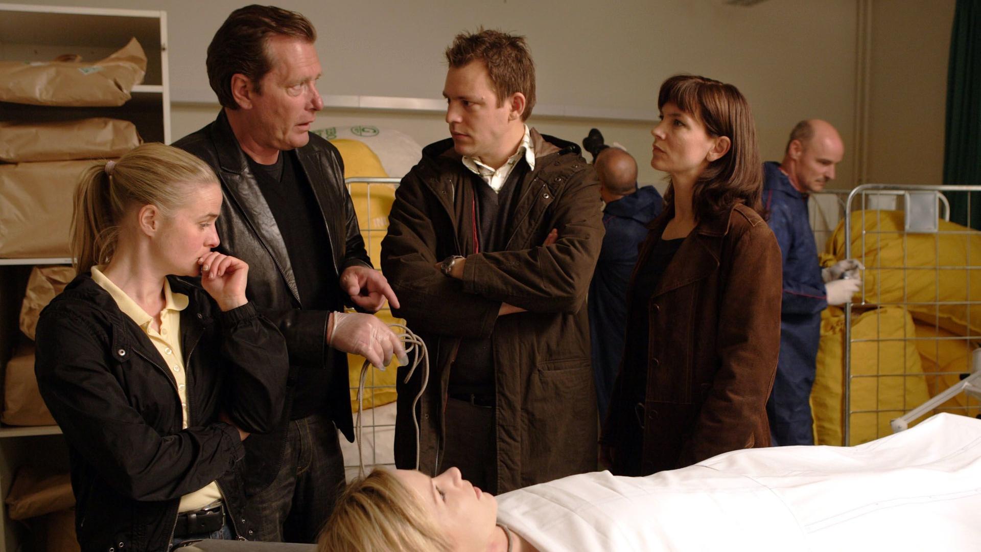 Zwei Männer und zwei Frauen stehen zusammen vor einer weiblichen Leiche, die auf einem Untersuchungstisch liegt und mit einem weißen Tuch bis zum Hals bedeckt ist.