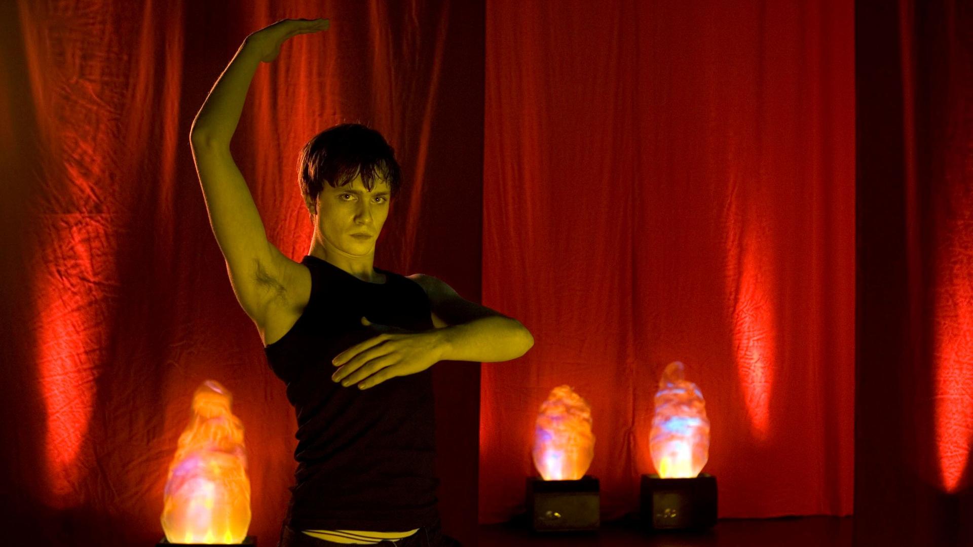 Ein junger Mann in tanzender Pose vor einem Hintergrund mit roten Vorhängen und rötlichen Stehleuchten
