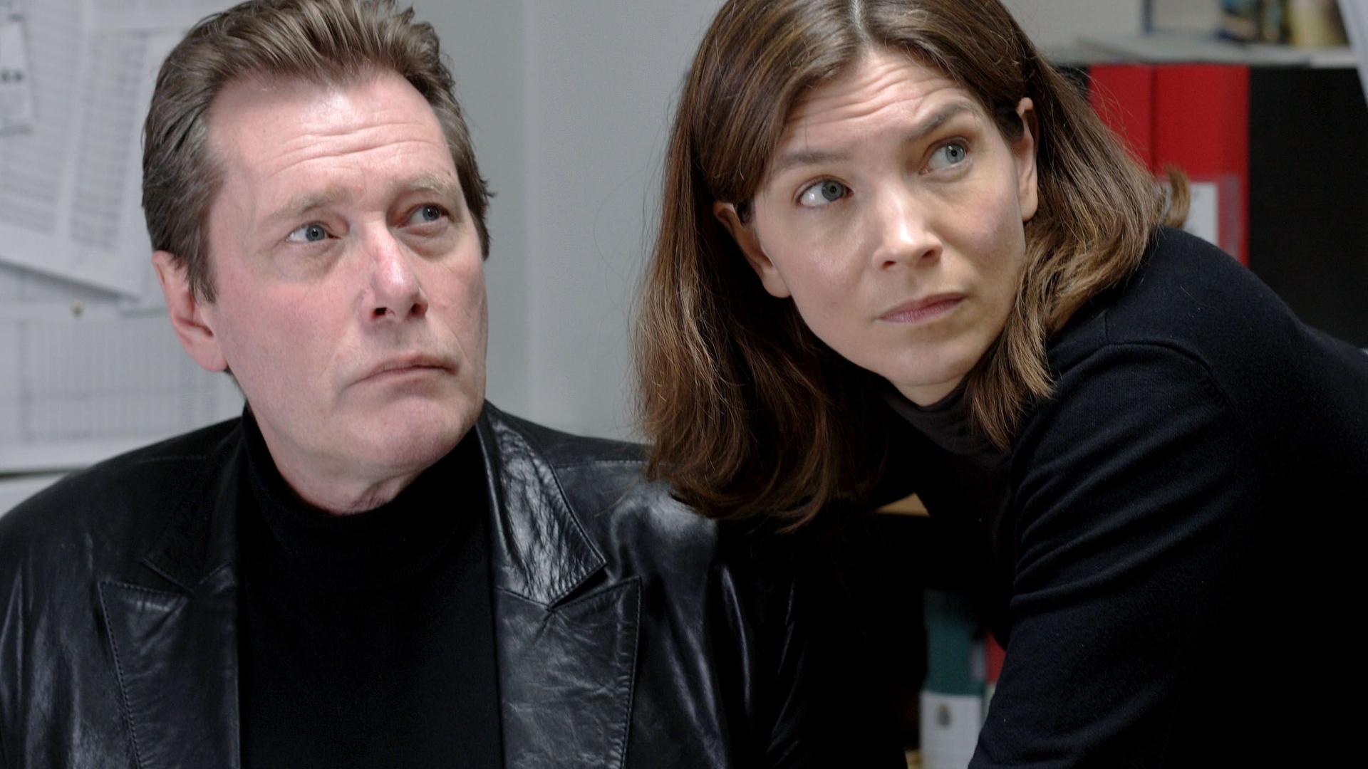 Eine Frau und ein Mann mittleren Alters schauen beide in dieselbe Richtung. Beide sind schwarz gekleidet.