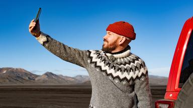 Ein Mann macht ein Selfie vor dem Hintergrund isländischer Landschaft.
