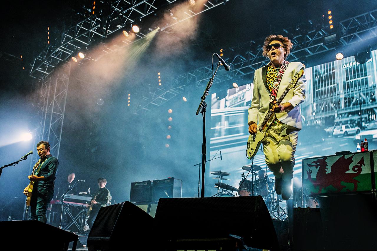 James Dean Bradfield springt auf der hell beleuchteten Bühne in die Luft. Er trägt einen Anzug in beige, ein buntes Hemd und hält die Bassgitarre in der Hand.