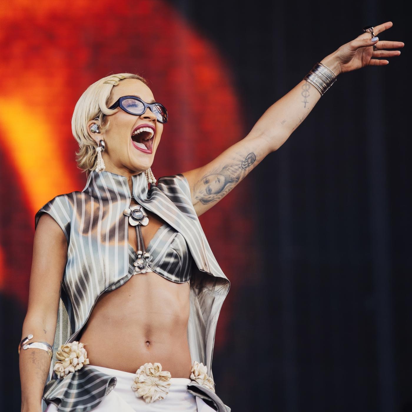 Rita Ora steht lächelnd auf der Bühne und hebt einen Arm in die Luft.
