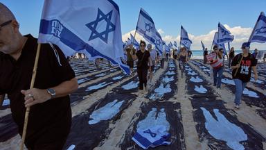 Menschen mit israelischen Flaggen laufen an einem Strand zwischen schwarzen Streifen im Sand, auf die weiß die Umrisse von Personen gemalt sind.