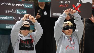Bei einer Demo halten zwei junge Menschen mit verbundenen Augen ihre gefesselten Hände hoch. Hinter ihnen stehen Menschen mit Plakaten, auf denen etwas in hebräischer Schrift geschrieben steht.
