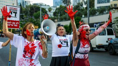 Drei Frauen, die sich mit roter Farbe beschmiert haben, laufen schreiend, mit hocherhobenen Händen bei einer Demonstration auf der Straße.