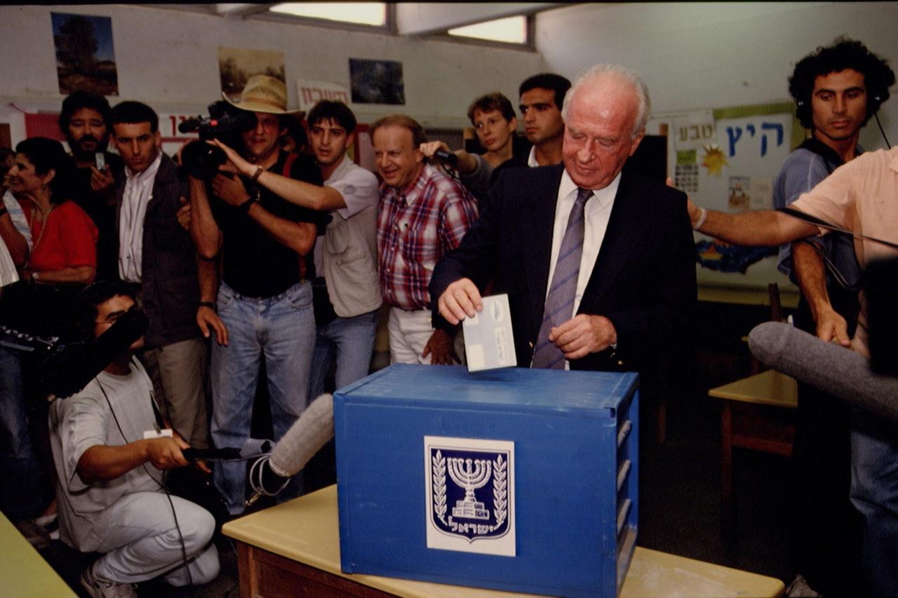 Jitzhak Rabin steht in Anzug und Krawatte hinter einer blauen Box und wirft ein gefaltetes Blatt hinein. Um ihn herum stehen zahlreiche Menschen, teilweise mit Kameras und Mikrophonen.