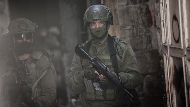 Nahaufnahme zweier Soldaten in grüner Kampfmontur mit vermummten Gesichtern und Gewehren gehen durch eine dunkle Straße.