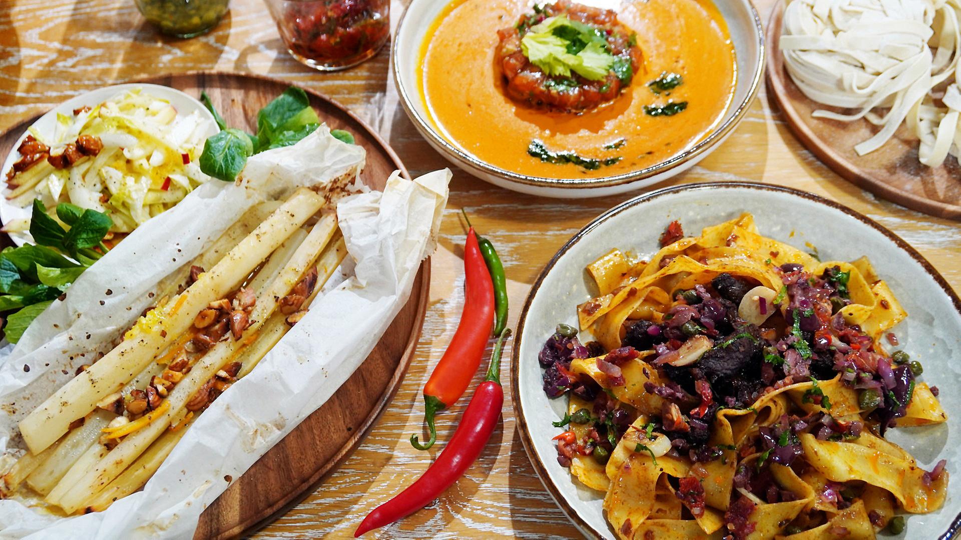 Ein Teller Currysuppe, Schwarzwurzeln in Pergament und Ragout vom Rind mit Pappardelle auf einem Tisch angerichtet.