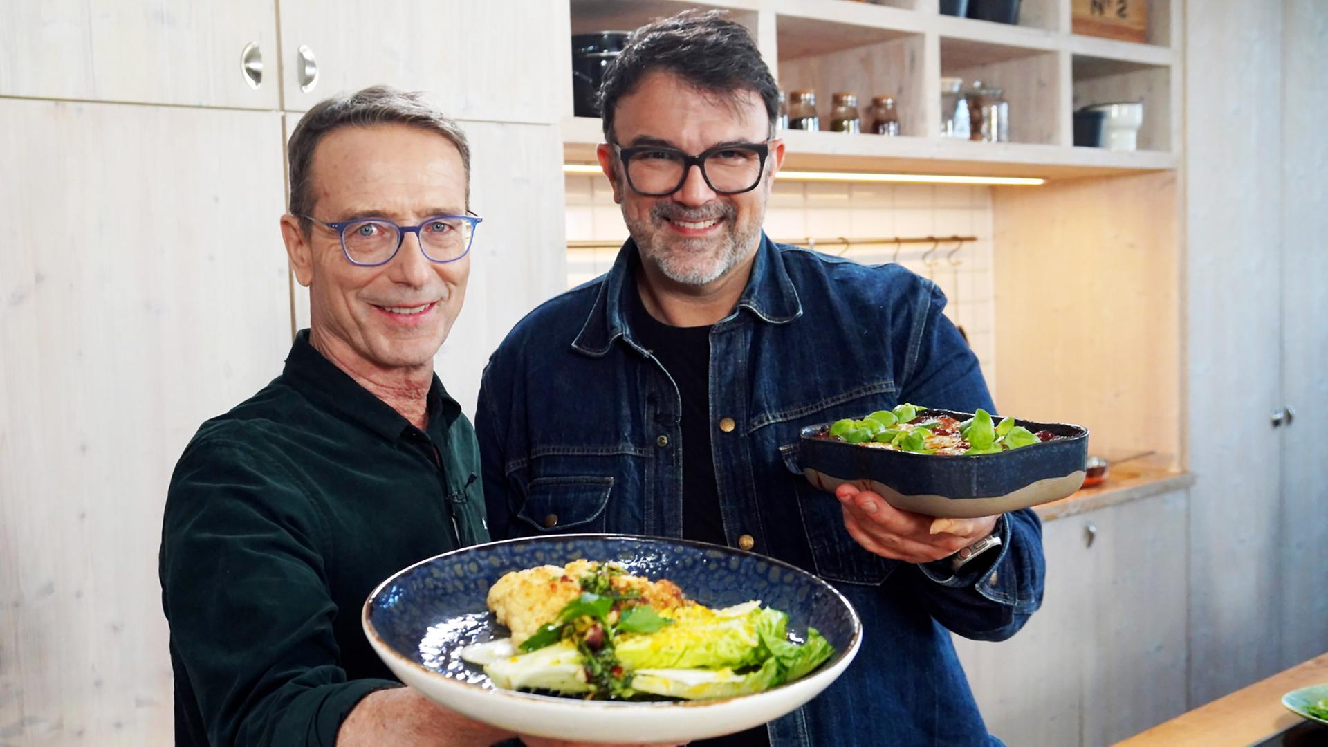 Tarik Rose und Dr. Matthias Riedl stehen in einer Küche und halten zwei Teller mit Ofengerichten in die Kamera.