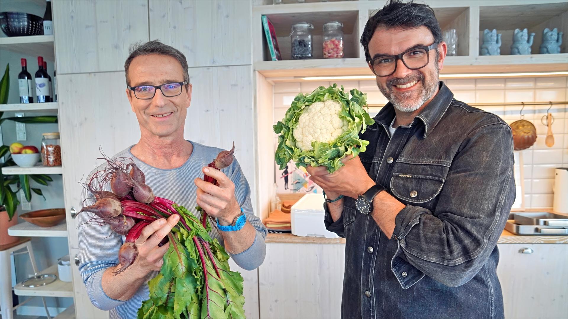 Fernsehkoch Tarik Rose (rechts) und Ernährungsmediziner Dr. Matthias Riedl stehen in der Küche und halten frische Rote Bete und einen Blumenkohl in die Kamera.
