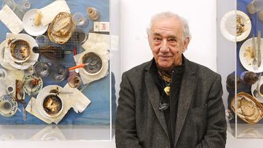 Der Künstler Daniel Spoerri steht zwischen zwei seiner Kunstwerken: der EatArt. Er trägt ein dunklesJackett und ein schwarzes Hemd. Er hält die Hände zusammen vor seinem Bauch. Auf seinen Bildern sind Kaffeetassen, Brot, Aufstriche und Servietten auf einem Tisch zu sehen.