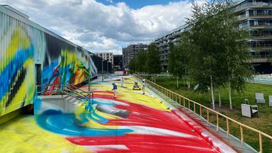 Vor einer Lagehalle stehen Menschen in Schutzanzügen und besprühen die Fassade sowie die Grünflächen darum herum mit bunten Farben.