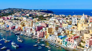Blick auf die Küste der italienischen Insel Procida mit dicht aneinandergereihten Häusern in Pastellfarben. Im Hafen liegen zahlreiche kleine Boote, dahinter erstreckt sich das Meer.