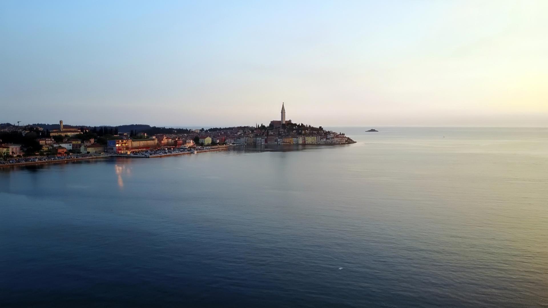 Die Küstenstadt Rovinj in Istrien, Kroatien, bei Sonnenuntergang. Die Stadt erhebt sich auf einer Halbinsel mit engen, pastellfarbenen Häusern und einem markanten Kirchturm in der Mitte.