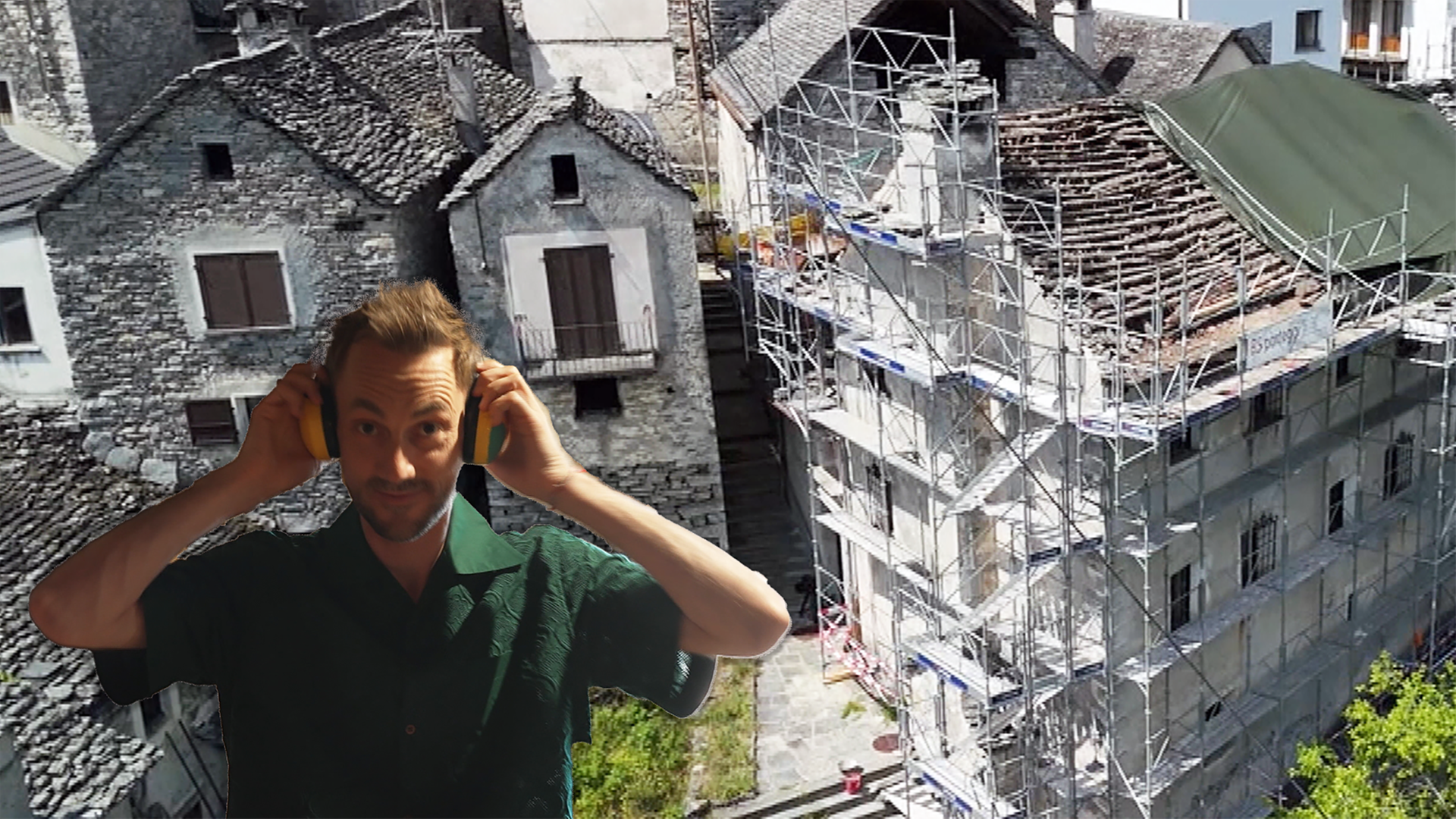 Ein Moderator Steht vor einem in Gerüste eingedecktes Haus mit Ohrenschutz.