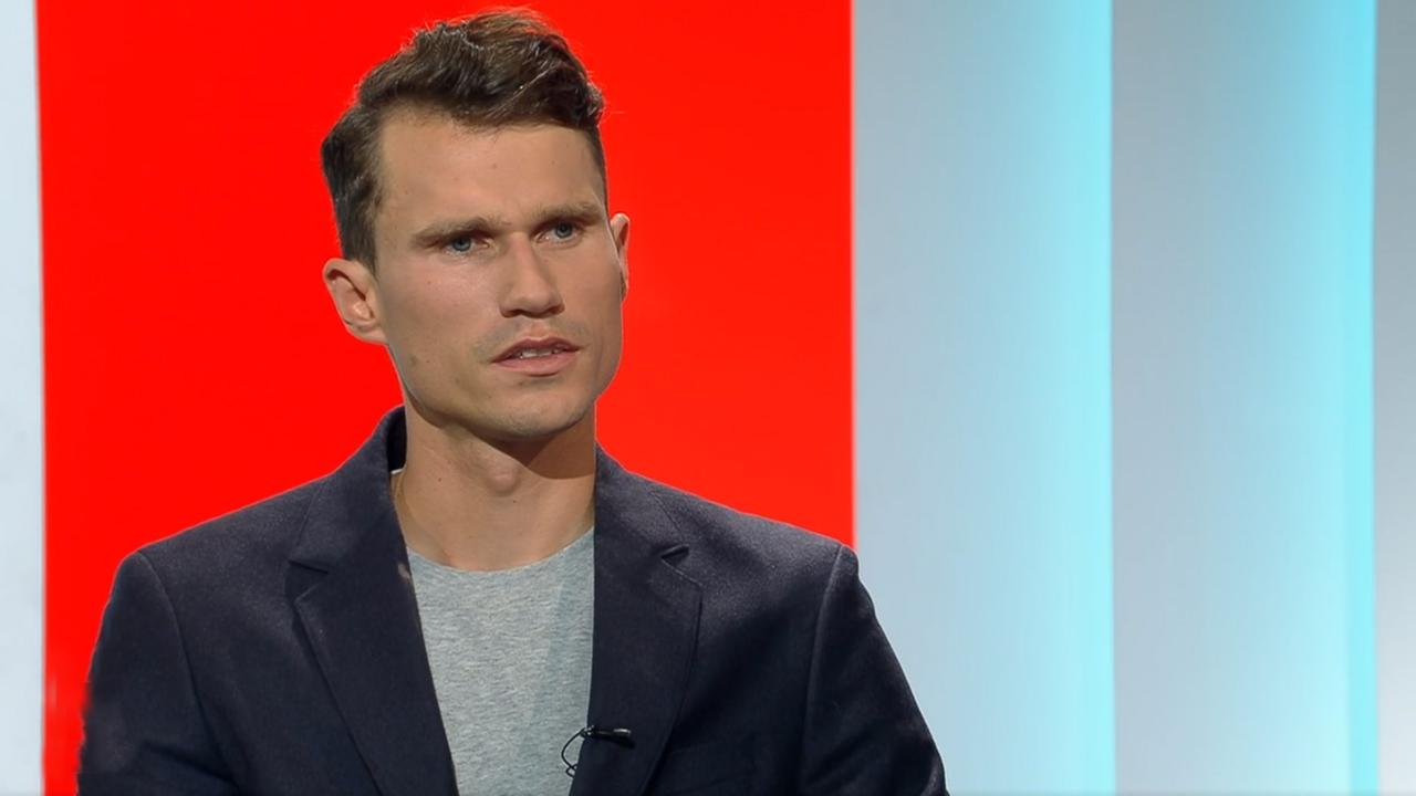 Ein Mann mit braunen Haaren und dunkelblauem Sacko sitzt vor einem roten Hintergrund. 