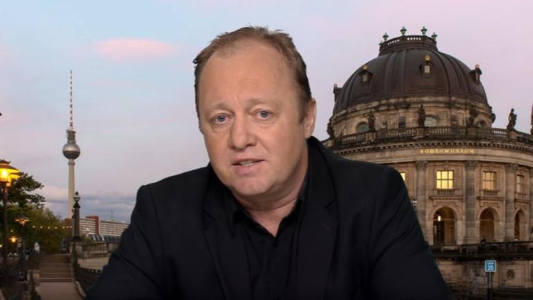 Ein Mann mit schwarzem Hemd sitzt vor einem Hintergrund der Stadt Berlin.