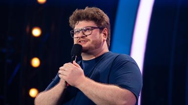 Jan Philipp Zymny steht in der Mitte des Bildes. Er hält mit beiden Händen das Mikrofon fest. Der Hintergrund ist blau beleuchtet und er trägt ein dunkelblaues T-Shirt.