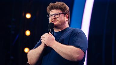 Jan Philipp Zymny steht in der Mitte des Bildes. Er hält mit beiden Händen das Mikrofon fest. Der Hintergrund ist blau beleuchtet und er trägt ein dunkelblaues T-Shirt.