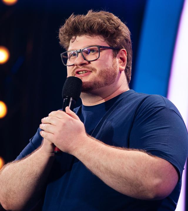 Jan Philipp Zymny steht in der Mitte des Bildes. Er hält mit beiden Händen das Mikrofon fest. Der Hintergrund ist blau beleuchtet und er trägt ein dunkelblaues T-Shirt.