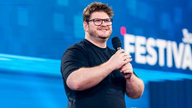 Jan Philipp Zymny steht in der Mitte des Bildes. Er lacht und hält sein Mikrofon mit beiden Händen fest. Das Licht auf der Bühne ist blau. Er trägt ein schwarzes Oberteil.