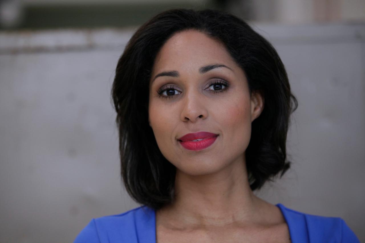 Ein Porträt einer Frau mit einem freundlichen Gesichtsausdruck. Die Komposition des Bildes ist einfach und konzentriert sich auf das Gesicht der Frau.  Der Blickwinkel ist direkt und frontal. Die Frau ist zentriert und nimmt den größten Teil des Bildfelds ein.  Der Hintergrund ist ein hellgraues, unstrukturiertes Muster.