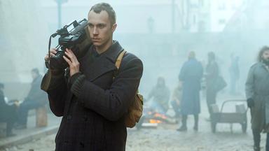 Ein junger Mann mit kurzen Haaren, Schnurrbart, grauem Mantel und Rucksack trägt eine Kamera auf der rechten Schulter. Er schaut resigniert, im Hintergrund sieht man unscharf einige Menschen an einem Lagerfeuer in der Stadt.