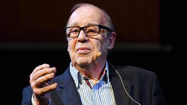 Mann im Anzug mit schwarzer Brille und angestecktem Mikrofon an der Wange.