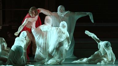 Bühnenszene: Ein Mann mit nacktem Oberkörper mit rotem Umhang bewegt sich zwischen in weiße Lanken gehüllte gespenstischen Gestalten.