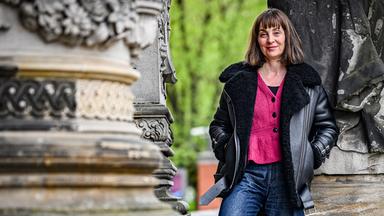 Eine Frau mit schulterlangen Haaren, bekleidet mit einer dicken Lederjacke, Jeans und pinkfarbener Strickjacke, posiert zwischen den dicken Säulen eines historischen Gebäudes für das Foto