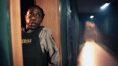 Tabby (Nikki Amuka-Bird) ist angsterfüllt und versteckt sich hinter einem Türrahmen. Der Gang neben ihr ist leer.