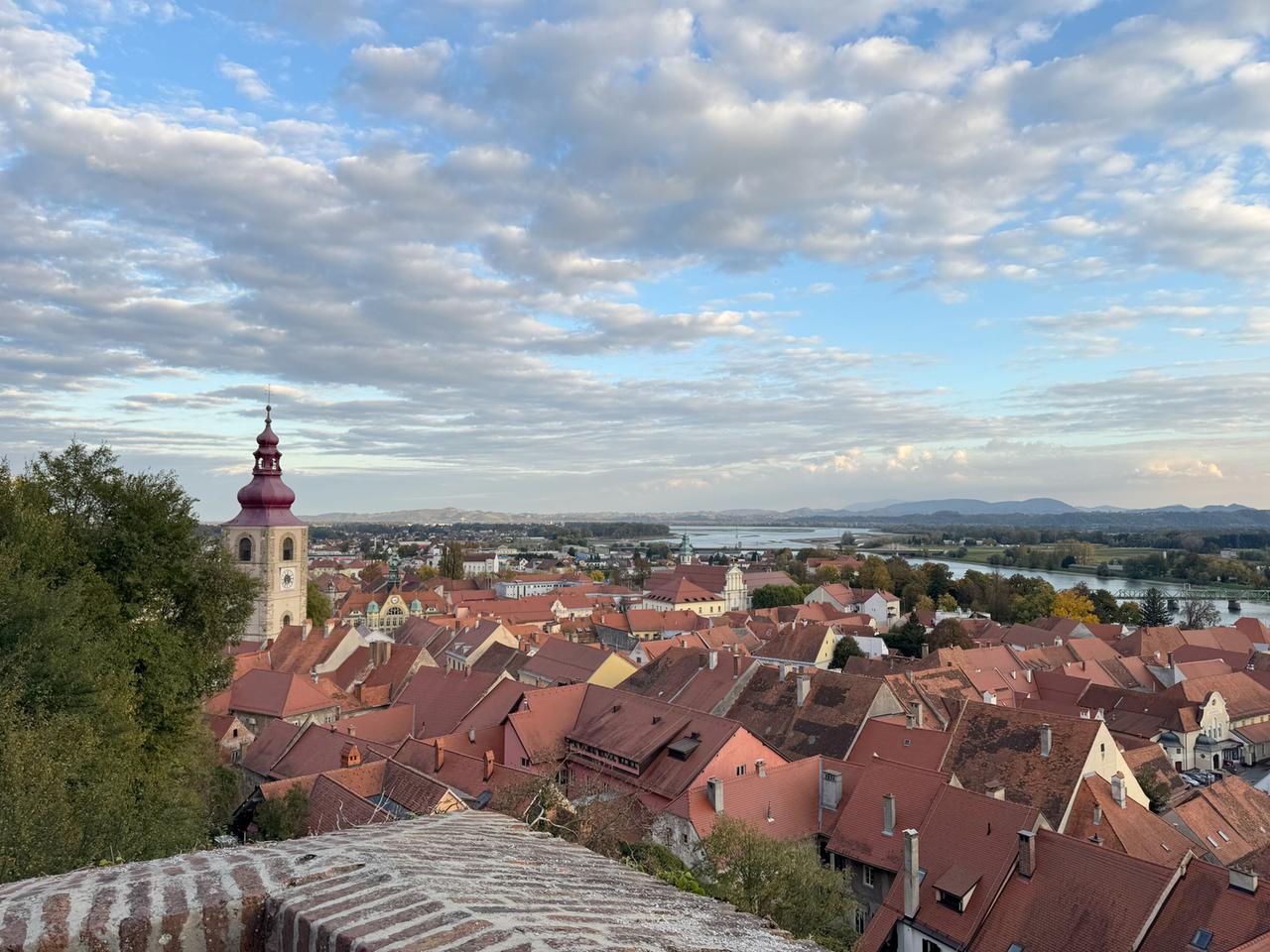 Das Bild zeigt eine panoramatische Aussicht auf die Stadt Ptuj (Pettau) in Slowenien. Im Vordergrund sind viele Dächer von Gebäuden sichtbar, die überwiegend rot geziegt sind. Daneben sind einige grüne Bäume zu erkennen. Ein markantes Gebäude mit einer hohen, spitzen Kirchturmhaube in Rot steht im Mittelpunkt des Bildes. Es hat eine Uhr an der Fassade und ist aus hellen Steinen errichtet.   Der Himmel ist teilweise bewölkt mit einer Mischung aus Wolken und blauen Abschnitten. Im Hintergrund erstreckt sich die Drau, ein Fluss, und es sind sanfte Hügel und Berge zu sehen. Dieses Landschaftsbild vermittelt ein Gefühl von Ruhe und landschaftlicher Schönheit, während sich die Architektur der Stadt harmonisch in die Umgebung einfügt.